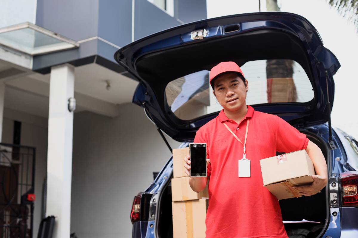 Courier stands in front of the trunk of a car filled with packages while holding a smartphone in his hand.